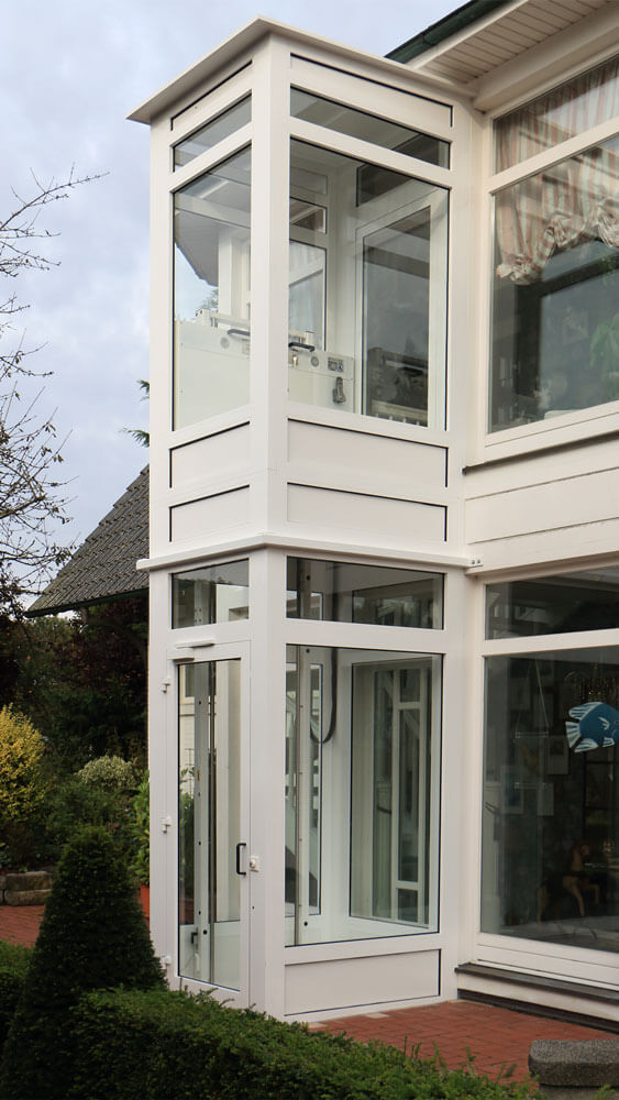 Weißer, verglaster Außenaufzug mit zwei Haltestellen, angebracht an einem Wohnhaus. Der Aufzug ermöglicht barrierefreien Zugang zwischen Erdgeschoss und Obergeschoss und fügt sich optisch in die moderne Architektur des Gebäudes ein.