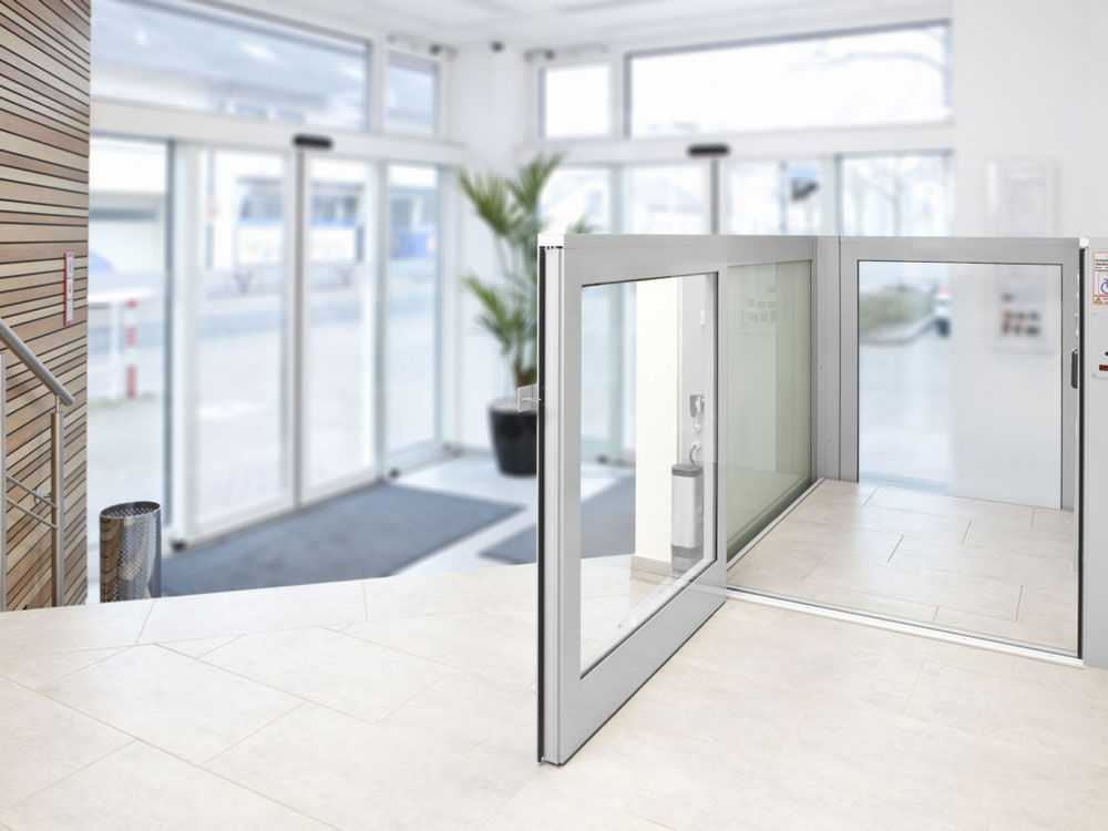 Barrierefreier Eingangsbereich mit automatischen Schiebetüren, gläsernem Aufzug für Rollstuhlfahrer, großen Fensterflächen und heller Innenarchitektur mit weißen Wänden und Fliesenboden – ideal für öffentliche Gebäude oder moderne Bürokomplexe.