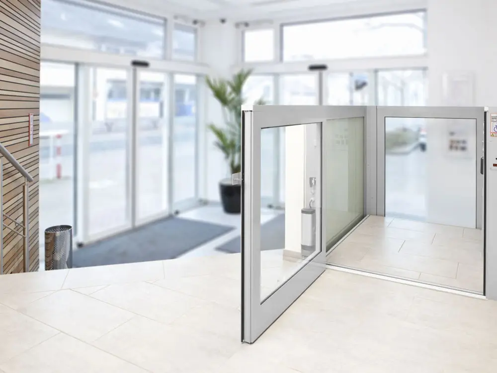 Barrierefreier Eingangsbereich mit automatischen Schiebetüren, gläsernem Aufzug für Rollstuhlfahrer, großen Fensterflächen und heller Innenarchitektur mit weißen Wänden und Fliesenboden – ideal für öffentliche Gebäude oder moderne Bürokomplexe.