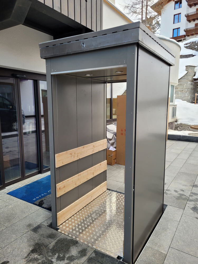 Unterflur-Liftsystem mit geschlossenem Metallgehäuse, Holzverkleidung im Innenbereich und rutschfester Bodenplatte, installiert im Außenbereich eines Gebäudes in winterlicher Umgebung – barrierefreie Lösung für Personen- oder Lastentransport bei kaltem Klima.