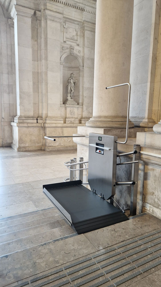 GTLE Plattform-Treppenlift am Fuß einer Steintreppe in einem Gebäude mit klassischer Architektur, ausgestattet mit Sicherheitsbügeln und Bedienfeld – barrierefreie Zugangslösung für Personen mit eingeschränkter Mobilität in historisch gestalteten öffentlichen Räumen.