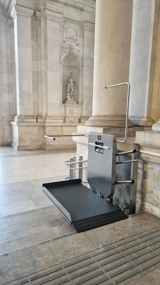 GTLE Plattform-Treppenlift am Fuß einer Steintreppe in einem Gebäude mit klassischer Architektur, ausgestattet mit Sicherheitsbügeln und Bedienfeld – barrierefreie Zugangslösung für Personen mit eingeschränkter Mobilität in historisch gestalteten öffentlichen Räumen.