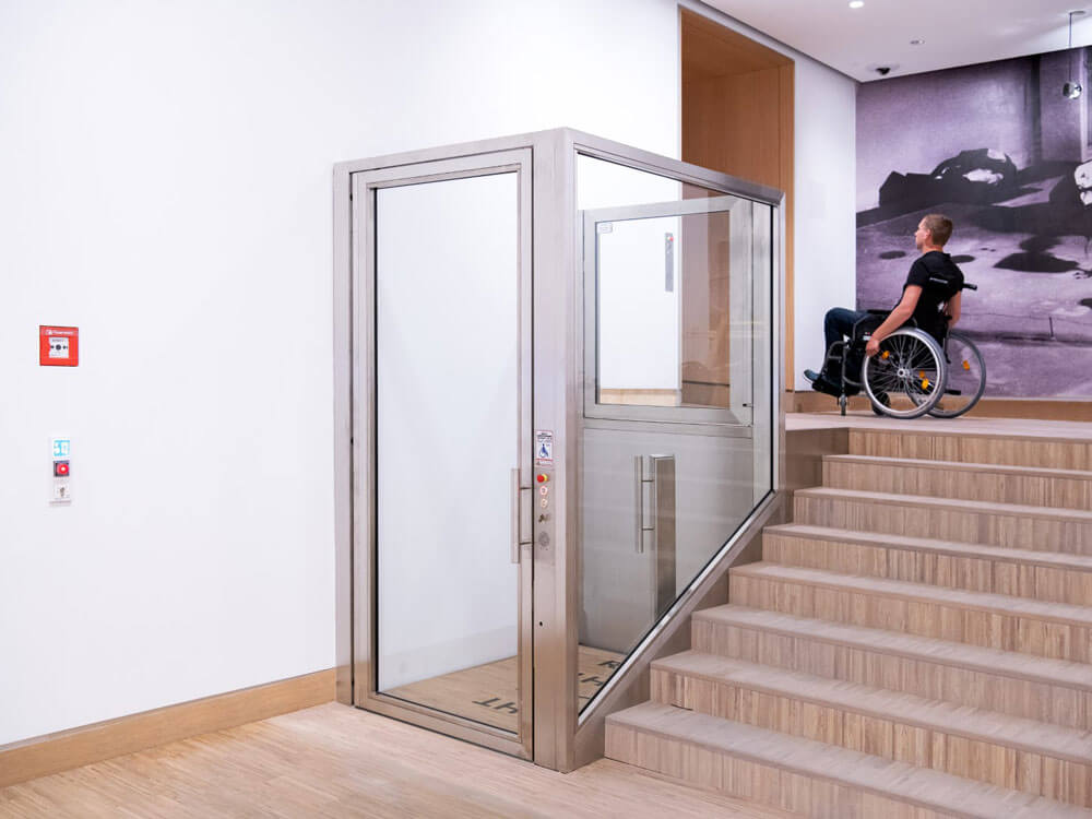 Barrierefreies Hebebühnen-System mit gläserner Einhausung und seitlichem Bedienpanel, installiert neben einer Treppe in einem modernen Innenraum – ermöglicht rollstuhlgerechten Zugang zwischen Ebenen, dargestellt mit einer Person im Rollstuhl am oberen Treppenabsatz.