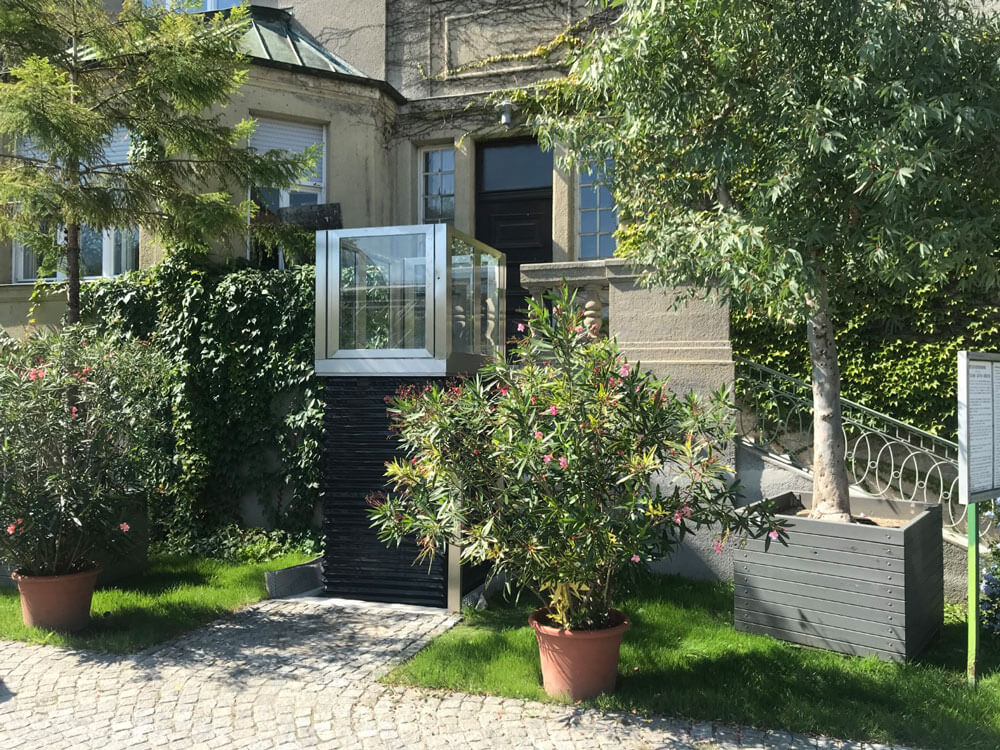 Hebebühne mit gläserner Einhausung und schwarzer Plattformbasis, installiert im Außenbereich einer Villa mit Steinfassade und großen Fenstern – barrierefreie Hebelösung für Personen oder Lasten, eingebettet in eine stilvolle und begrünte Umgebung.