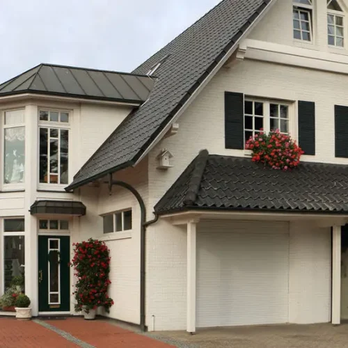 Zweistöckiges Wohnhaus mit weißer Fassade und dunkelgrauem Dach. Große Fenster mit schwarzen Fensterläden und roten Blumenkästen. Rechts befindet sich eine Garage mit geschlossener weißer Tür. Der Hauseingang hat eine grüne Tür mit seitlichen Glaspaneelen. Vor dem Haus liegt eine gepflasterte Einfahrt mit Pflanzen und Sträuchern.