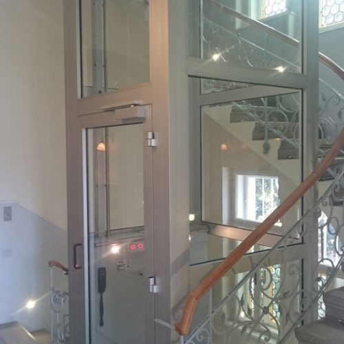 Verglaster Aufzug mit Metallrahmen, eingebaut in ein Treppenhaus mit dekorativen Metallgeländern und Holzhandläufen. Der Aufzug bietet barrierefreien Zugang und verbindet moderne Technik mit klassischer Architektur.