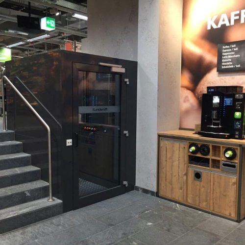 Innenbereich eines Geschäfts mit einem kleinen Aufzug mit Glastür und der Beschriftung ‚Kundelift‘ links neben einer Treppe mit Metallgeländer. Rechts befindet sich eine Kaffeestation mit Holzverkleidung, Kaffeemaschinen und einer großen Preistafel mit verschiedenen Kaffeesorten.