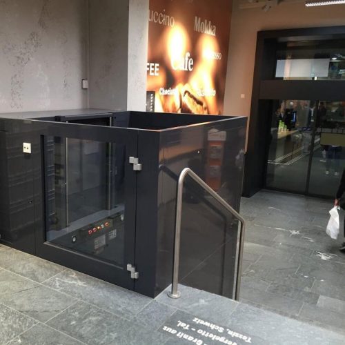 Blick nach unten in einen verglasten Aufzugsschacht mit transparenten Türen und Wänden. Sichtbar sind Führungsschienen und Kabel entlang der Seiten. Teile des Gebäudes mit Wanddekorationen und Bildern sind außerhalb des Schachts erkennbar.