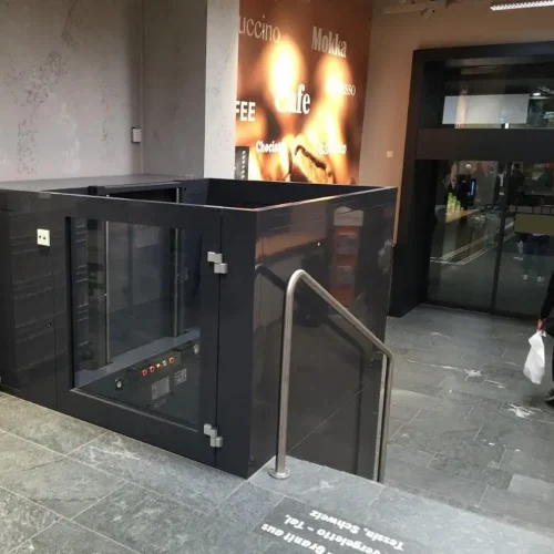 Blick nach unten in einen verglasten Aufzugsschacht mit transparenten Türen und Wänden. Sichtbar sind Führungsschienen und Kabel entlang der Seiten. Teile des Gebäudes mit Wanddekorationen und Bildern sind außerhalb des Schachts erkennbar.