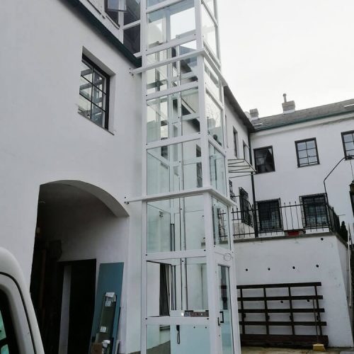 Außenaufzug mit verglasten Wänden und weißem Rahmen, installiert im Innenhof eines Gebäudes. Der Aufzug ermöglicht barrierefreien Zugang zu mehreren Etagen. Umgeben von weißen Fassaden mit schwarzen Fensterrahmen und sichtbaren Baustellenelementen.