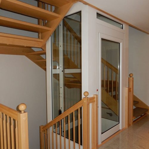 Moderne Innentreppe mit Holzstufen und Holzgeländer, die sich um einen verglasten Wohnhausaufzug mit weißem Rahmen windet. Der Aufzug ermöglicht barrierefreien Zugang zu mehreren Etagen innerhalb des Hauses.