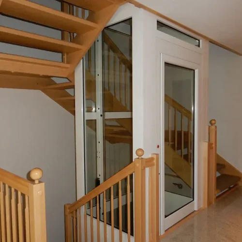 Moderne Innentreppe mit Holzstufen und Holzgeländer, die sich um einen verglasten Wohnhausaufzug mit weißem Rahmen windet. Der Aufzug ermöglicht barrierefreien Zugang zu mehreren Etagen innerhalb des Hauses.