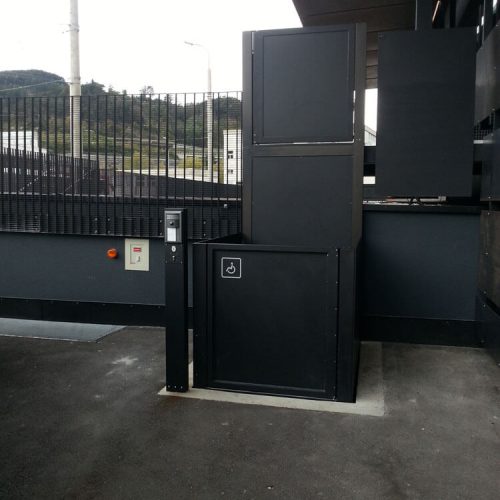 Schwarzer Plattformlift im Außenbereich mit Rollstuhlsymbol, installiert neben einem Zaun. Der Lift dient der barrierefreien Überwindung von Höhenunterschieden für Personen mit Mobilitätseinschränkungen.