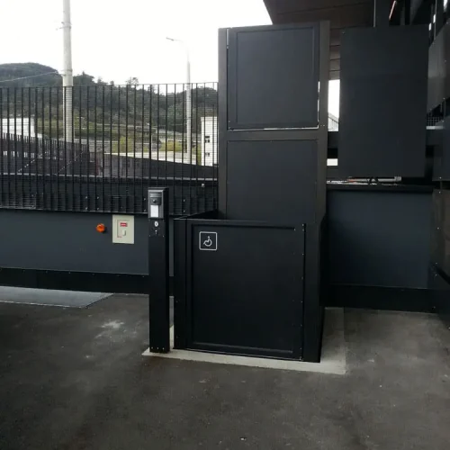 Schwarzer Plattformlift im Außenbereich mit Rollstuhlsymbol, installiert neben einem Zaun. Der Lift dient der barrierefreien Überwindung von Höhenunterschieden für Personen mit Mobilitätseinschränkungen.