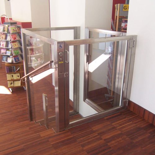 Kleiner Aufzug mit Glaswänden und Edelstahlrahmen in einem öffentlichen Innenraum, vermutlich einer Bibliothek oder Buchhandlung. Der Aufzug befindet sich neben einer Treppe und ermöglicht barrierefreien Zugang für Personen mit Mobilitätseinschränkungen. Im Hintergrund sind Holzfußboden und Bücherregale sichtbar.