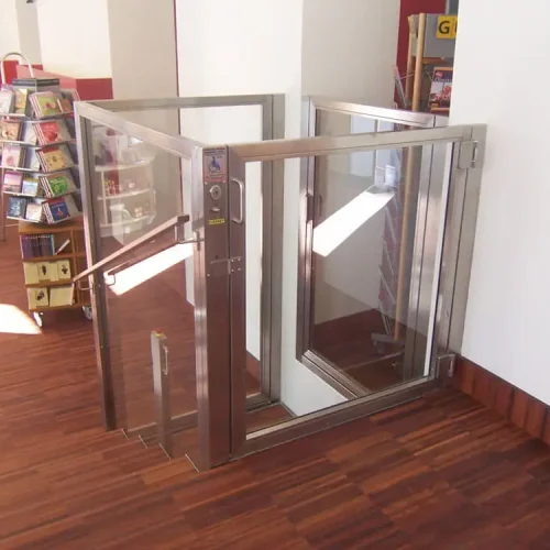 Kleiner Aufzug mit Glaswänden und Edelstahlrahmen in einem öffentlichen Innenraum, vermutlich einer Bibliothek oder Buchhandlung. Der Aufzug befindet sich neben einer Treppe und ermöglicht barrierefreien Zugang für Personen mit Mobilitätseinschränkungen. Im Hintergrund sind Holzfußboden und Bücherregale sichtbar.