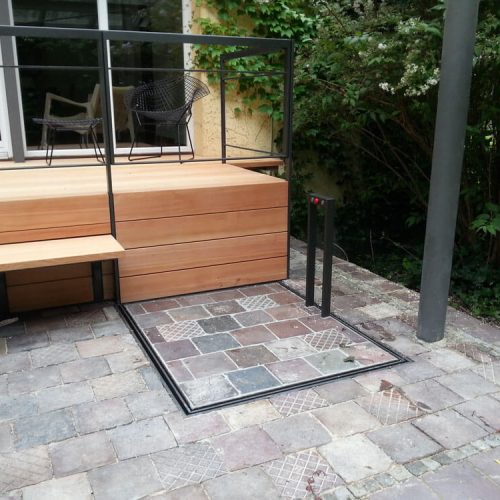 Unterflur-Liftsystem neben einer erhöhten Holzterrasse mit Treppen und schwarzem Geländer, eingebaut in einen gepflasterten Außenbereich mit Steuerungseinheit – barrierefreie Zugangslösung für Personen mit eingeschränkter Mobilität in einem begrünten Wohnumfeld.
