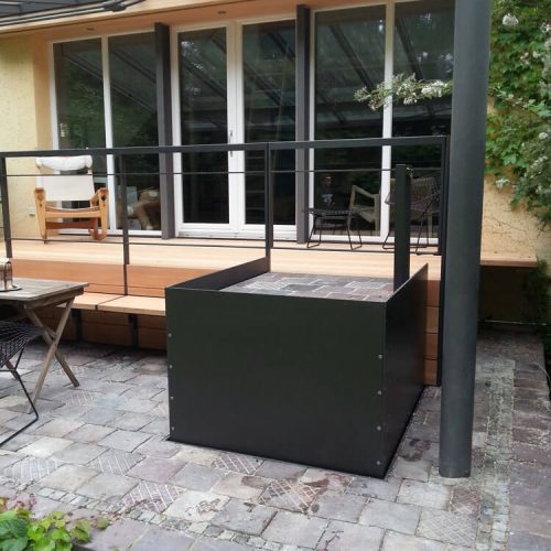 Unterflur-Liftsystem mit schwarzem Metallrahmen und integrierter Plattform, eingebaut in eine moderne Holzterrasse mit Glasüberdachung; umgeben von Gartenmöbeln und Begrünung – barrierefreie Zugangslösung für private Wohnbereiche mit stilvoller Außengestaltung.