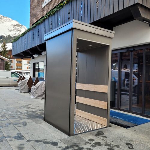 Unterflur-Liftsystem vor dem ARLBERG CLUB HOTEL mit metallischer Konstruktion und Holzverkleidung im Innenbereich, installiert auf einem gepflasterten Außenbereich in alpiner Umgebung – barrierefreie Zugangslösung für Hotelgäste in winterlichem Klima mit Blick auf schneebedeckte Berge.