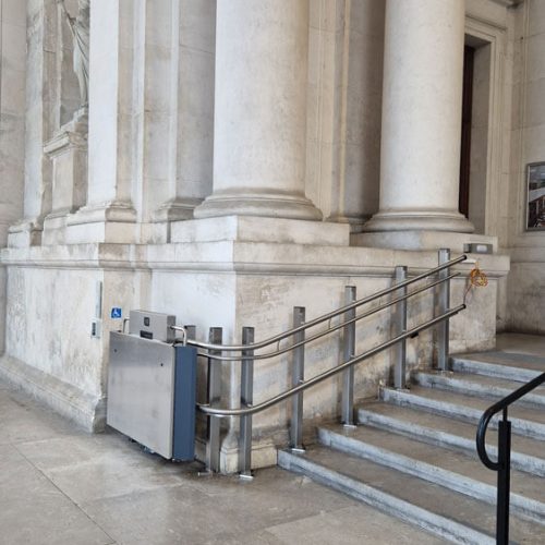 GTLE Plattform-Treppenlift neben einer Treppe mit Metallgeländer am Eingang eines Gebäudes mit weißen Steinsäulen – barrierefreie Zugangslösung mit Rollstuhlsymbol für Personen mit eingeschränkter Mobilität im öffentlichen Raum.