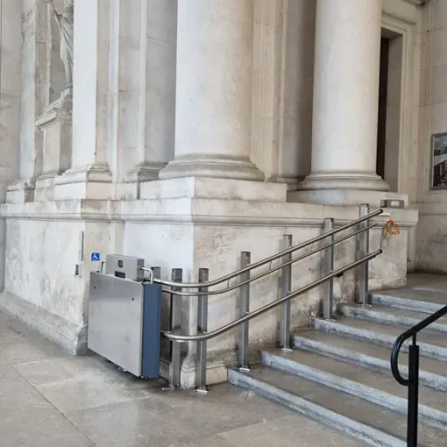 GTLE Plattform-Treppenlift neben einer Treppe mit Metallgeländer am Eingang eines Gebäudes mit weißen Steinsäulen – barrierefreie Zugangslösung mit Rollstuhlsymbol für Personen mit eingeschränkter Mobilität im öffentlichen Raum.