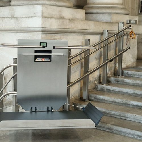 GTLE Plattform-Treppenlift in abgesenkter Position neben einer Treppe in einem öffentlichen Gebäude, ausgestattet mit Handläufen und einem Hinweisschild zur Barrierefreiheit – sichere und inklusive Zugangslösung für Personen mit Mobilitätseinschränkungen.