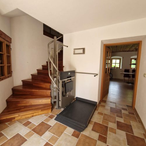 GTLE Plattform-Treppenlift am Fuß einer Holztreppe im Innenbereich eines Wohnhauses, mit seitlichem Bedienfeld und stabiler Plattform – barrierefreie Hebelösung für Personen mit eingeschränkter Mobilität zur sicheren Überwindung von Treppen im privaten Umfeld.