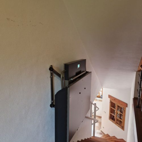 GTLE Plattform-Treppenlift im zusammengeklappten Zustand, montiert an einer Holztreppe im Innenbereich eines Wohnhauses – platzsparende und barrierefreie Lösung für Personen mit eingeschränkter Mobilität zur sicheren Überwindung von Treppen.