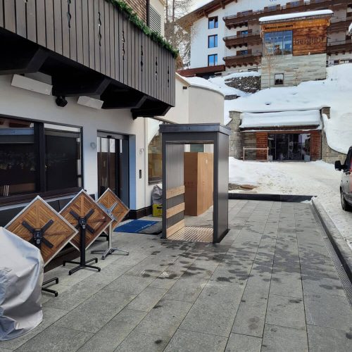 Unterflur-Liftsystem mit gläserner Einhausung im Außenbereich eines alpinen Hotelgebäudes; installiert auf gepflastertem Boden mit Anzeichen von Schneeschmelze, umgeben von modernen Fassaden und Holzverkleidungen – barrierefreie Zugangslösung für Personen oder Transport in winterlichen Regionen.