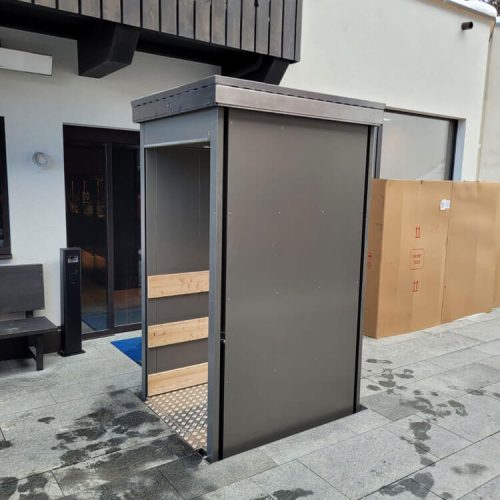 Seitliche Ansicht eines Unterflur-Liftsystems mit metallischer Konstruktion und rutschfester Bodenplatte, eingebaut im Außenbereich vor einem modernen Gebäude mit großen Fenstern – barrierefreie Lösung für den vertikalen Transport von Personen oder Lasten im urbanen Umfeld.