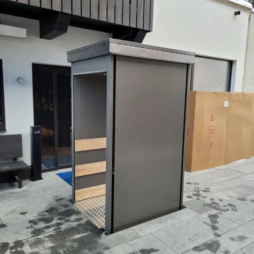 Seitliche Ansicht eines Unterflur-Liftsystems mit metallischer Konstruktion und rutschfester Bodenplatte, eingebaut im Außenbereich vor einem modernen Gebäude mit großen Fenstern – barrierefreie Lösung für den vertikalen Transport von Personen oder Lasten im urbanen Umfeld.
