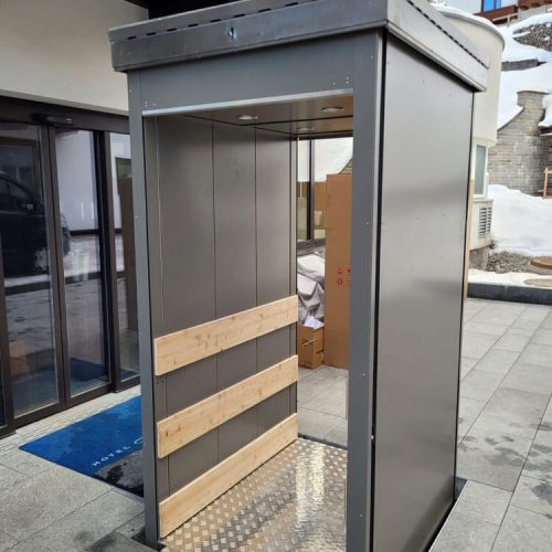 Außenaufzug mit offenem, metallischem Gehäuse und rutschfester Bodenplatte aus Riffelblech, ausgestattet mit Holzplanken zur Stabilisierung; installiert neben einem modernen Gebäude mit Glasfassade, geeignet für barrierefreien Zugang in kalten Klimazonen.