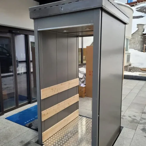 Außenaufzug mit offenem, metallischem Gehäuse und rutschfester Bodenplatte aus Riffelblech, ausgestattet mit Holzplanken zur Stabilisierung; installiert neben einem modernen Gebäude mit Glasfassade, geeignet für barrierefreien Zugang in kalten Klimazonen.