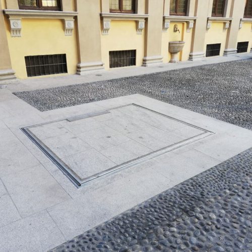 Rechteckige Unterflur-Klappe aus Metall, eingebettet in einen gepflasterten Hof mit umliegenden Steinplatten; im Hintergrund ein gelbes Gebäude mit klassischen Fassadenelementen, ein geparktes Auto und ein roter Hydrant – funktionale Lösung für Wartungs- oder Versorgungszugänge im urbanen Raum.