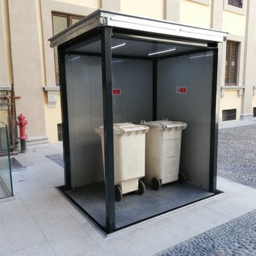 Unterflur-Mülltonnen-System mit zwei großen Abfallbehältern in einem modernen, wettergeschützten Metall- und Glasgehäuse auf einem gepflasterten Außenbereich nahe eines Gebäudes – saubere und organisierte Lösung für die Abfallentsorgung in öffentlichen oder gemeinschaftlich genutzten Räumen.