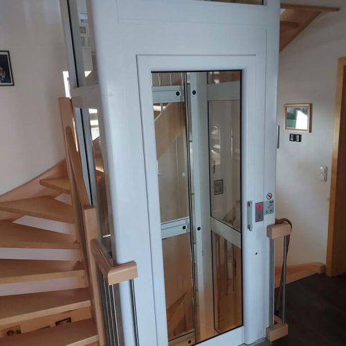 Kleiner Wohnhausaufzug mit weißem Rahmen und Glaspaneelen, installiert neben einer Holz-Wendeltreppe. Der Aufzug verfügt über ein Bedienfeld mit Tasten und Anzeigen auf der rechten Seite der Tür und ermöglicht barrierefreien Zugang zwischen Etagen.