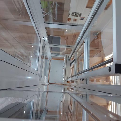 Blick von oben in einen verglasten Aufzugsschacht mit transparenten Wänden und Türen. Metallrahmen und Führungsschienen strukturieren die Konstruktion. Die Glasflächen reflektieren das Licht und geben den Blick bis zum Boden des Schachts frei.