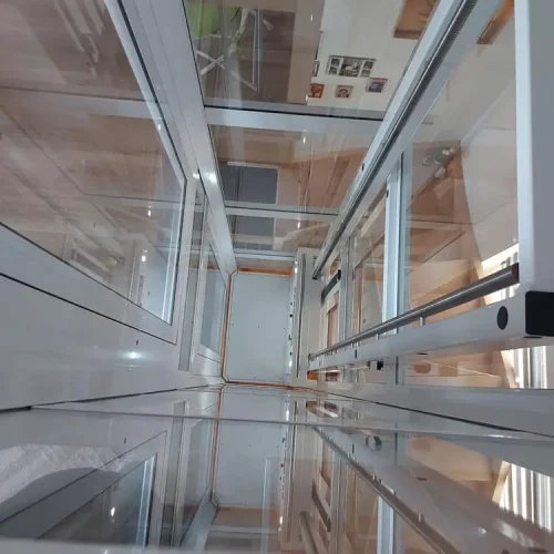 Blick von oben in einen verglasten Aufzugsschacht mit transparenten Wänden und Türen. Metallrahmen und Führungsschienen strukturieren die Konstruktion. Die Glasflächen reflektieren das Licht und geben den Blick bis zum Boden des Schachts frei.