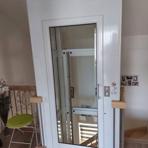 Kompakter Aufzug mit weißem Gehäuse und Glastür in einem Wohnhaus. Rechts neben der Tür befinden sich Bedientasten, ein Schlüsselschalter sowie Sicherheitshinweise. Der Aufzug steht neben einem kleinen Sitzbereich mit grünem Stuhl und Blumendekoration.