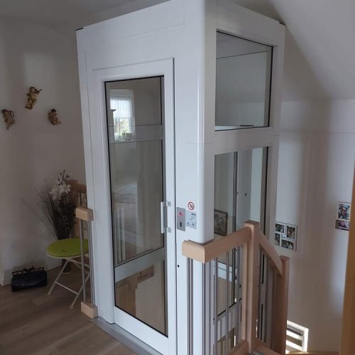 Wohnhaus mit modernem Aufzug mit weißem Rahmen und Glaspaneelen, installiert neben einer Holztreppe mit Metallgeländern. Der Aufzug ermöglicht barrierefreien Zugang zwischen Etagen. Im Raum befinden sich dekorative Elemente wie Wandbilder, ein kleiner grüner Tisch mit Stühlen und Wandornamente