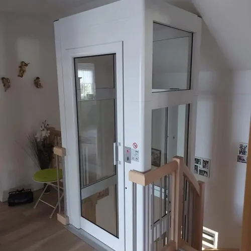 Wohnhaus mit modernem Aufzug mit weißem Rahmen und Glaspaneelen, installiert neben einer Holztreppe mit Metallgeländern. Der Aufzug ermöglicht barrierefreien Zugang zwischen Etagen. Im Raum befinden sich dekorative Elemente wie Wandbilder, ein kleiner grüner Tisch mit Stühlen und Wandornamente