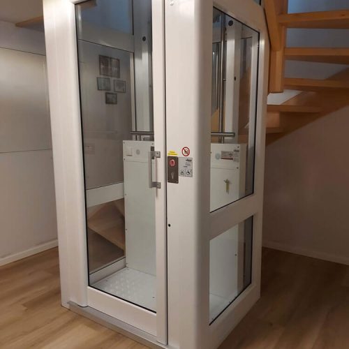 Kompakter, moderner Aufzug mit Glaspaneelen in einem Wohnhaus, installiert neben einer Holztreppe. Der Aufzug dient als barrierefreie Alternative zur Treppe und verfügt über ein Bedienfeld mit Tasten und Sicherheitshinweisen.