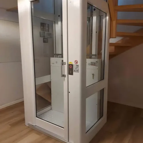 Kompakter, moderner Aufzug mit Glaspaneelen in einem Wohnhaus, installiert neben einer Holztreppe. Der Aufzug dient als barrierefreie Alternative zur Treppe und verfügt über ein Bedienfeld mit Tasten und Sicherheitshinweisen.