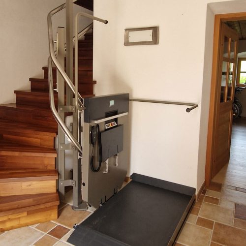 Hebebühne für barrierefreien Zugang im Innenbereich eines Wohnhauses, installiert neben einer Holztreppe mit seitlichem Bedienpanel – ermöglicht sicheren und komfortablen Etagenwechsel für Personen mit eingeschränkter Mobilität, umgeben von Fliesenboden und offenem Wohnbereich.